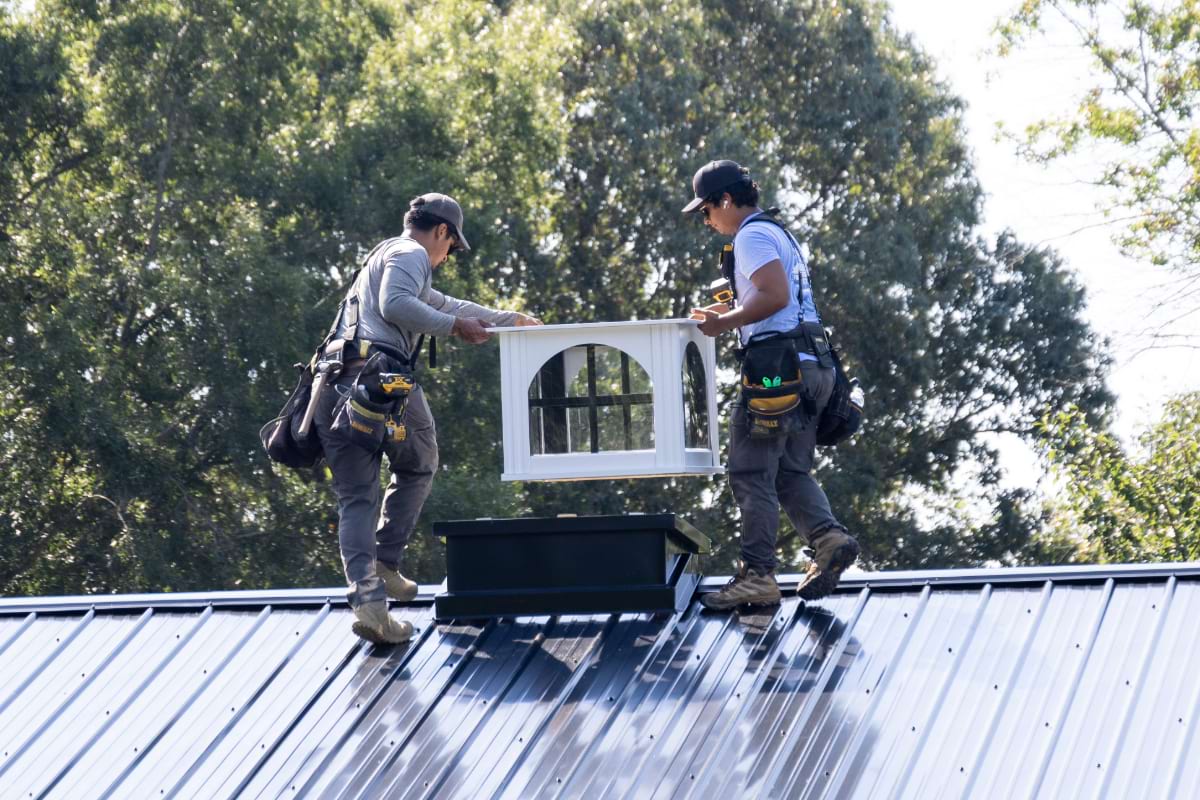 two blue steel manufacturing workers on top of a roof attaching a cupola to the roof